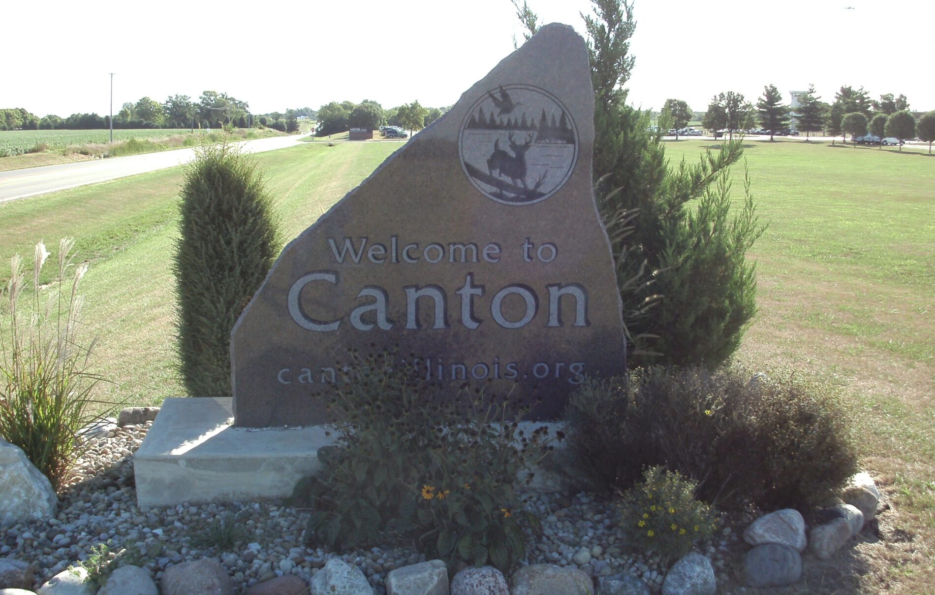 Large stone sign reads "Welcome to Canton" by a roadside, surrounded by grass, shrubs, and rocks.