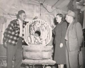 Three people stand around a large circular object with a lion illustration on it inside an indoor setting.