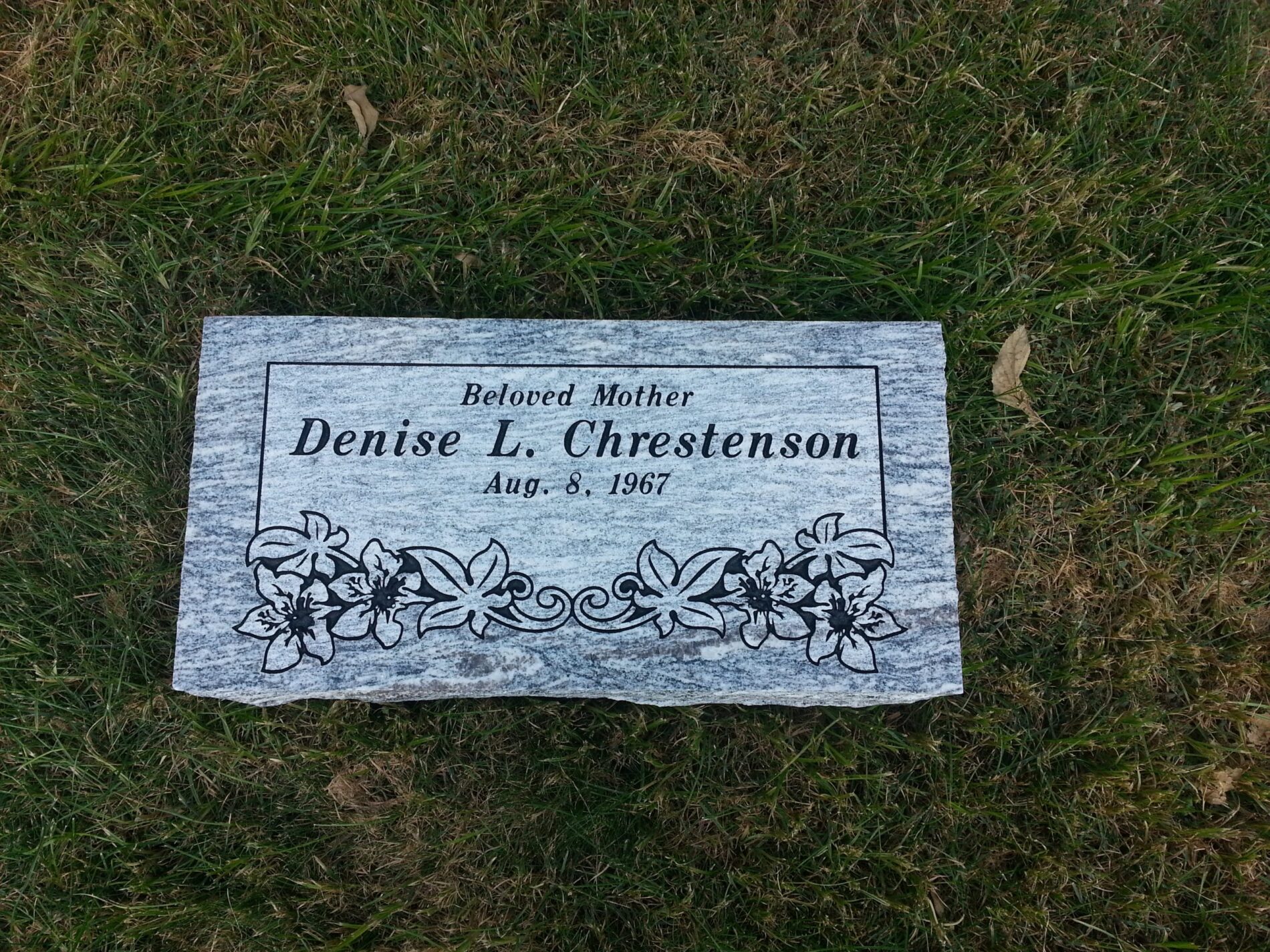 A gravestone reads "Beloved Mother Denise L. Chrestenson Aug. 8, 1967" with floral carvings, set on grass.