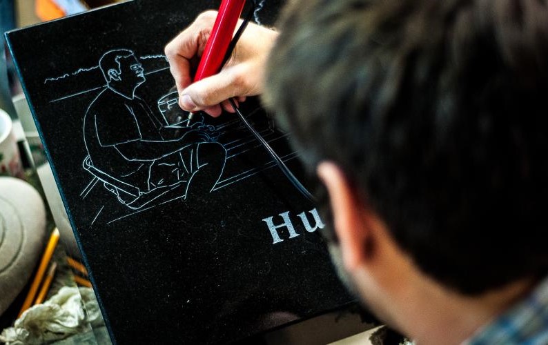 A person engraves an outline of a seated man on a black stone surface with a red tool.
