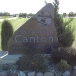 Large stone sign reads "Welcome to Canton" by a roadside, surrounded by grass, shrubs, and rocks.